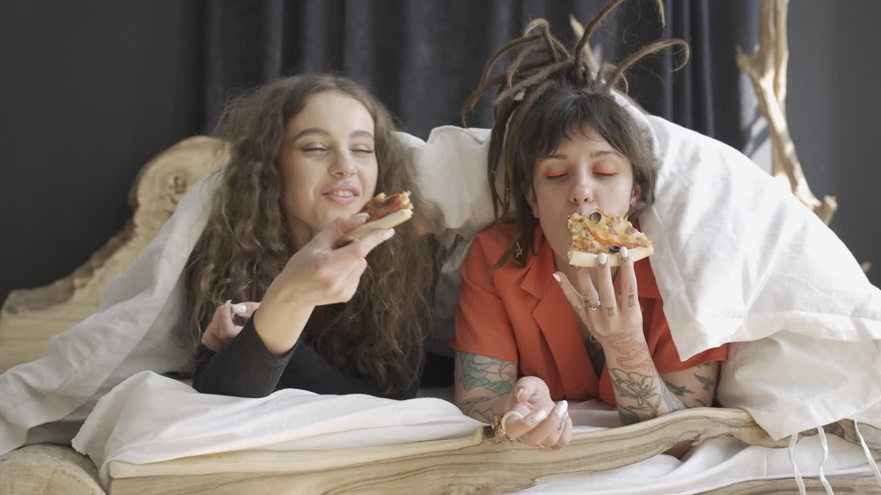 amigas jóvenes acostadas en la cama mientras hablan y comen pizza