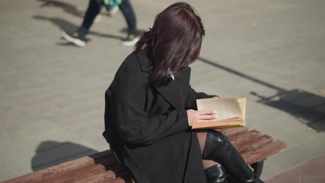 woman concentrating on reading her book with her hand on the page, hair cascading over her shoulder, long shadow cast on the ground, with blurred view of people passing behind