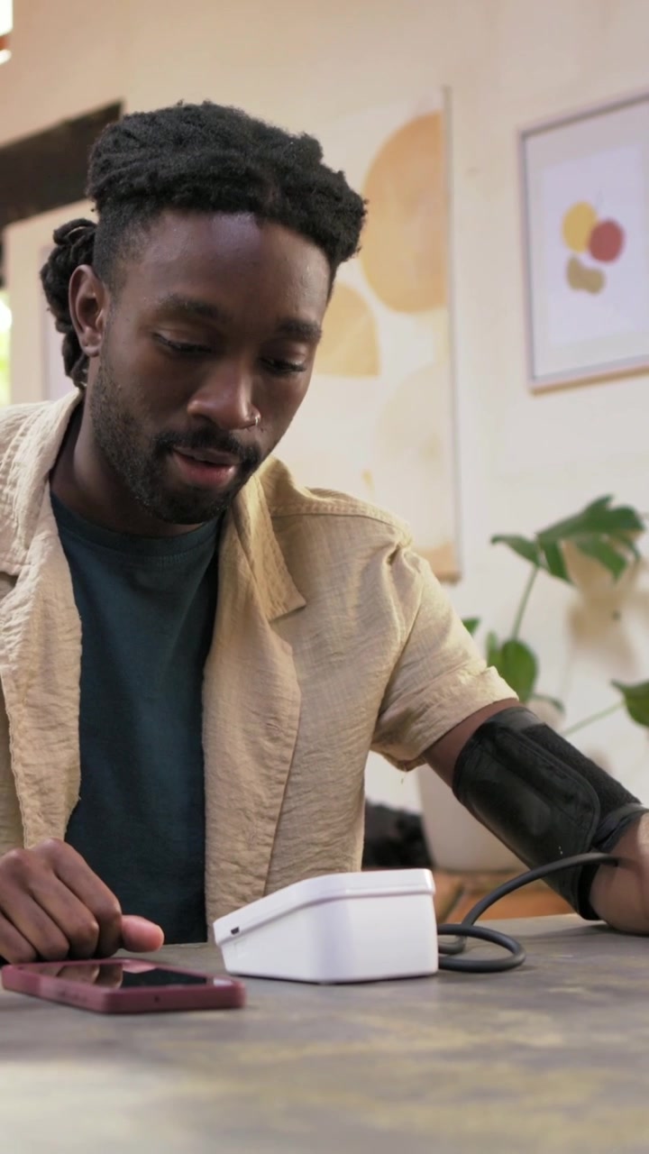 Vertical video: Taking BP, adult man positioning arm on table at home, tapping phone, starting cuff