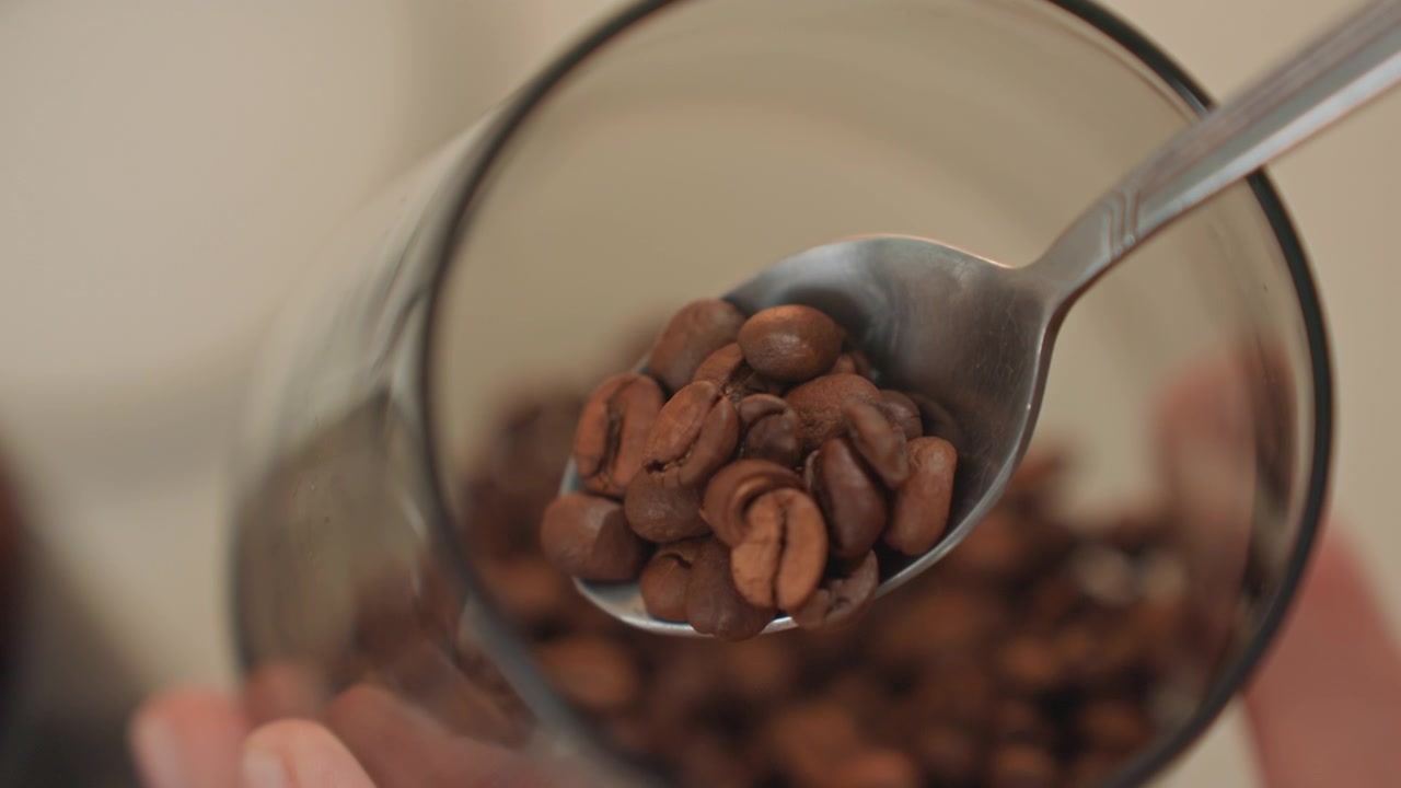 Cucchiaio che raccoglie chicchi di caffè da un barattolo di vetro, primo piano su un utensile metallico lucido che solleva una porzione misurata, mani ferme in una cucina studio, ritmo giocoso e atmosfera musicale energica, barista che prepara.