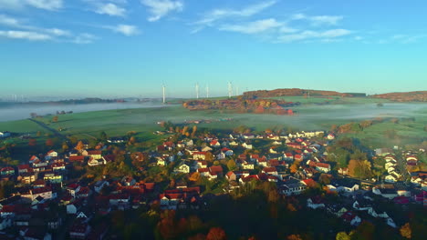 Vista-Aérea-Sobre-Una-Ciudad-Europea-Con-Fondo-De-Turbina-De-Viento,-Amanecer-Brumoso