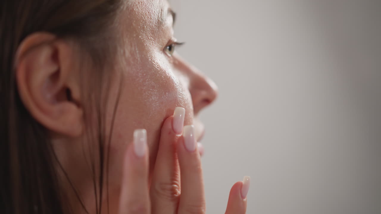 여성은 손가락 끝으로 얼굴 보습제를 깨뜨립니다. 어두운 머리카락을 가진 젊은 여성은 화장실에서 투명한 보습 크림으로 얼굴을 마사지합니다.