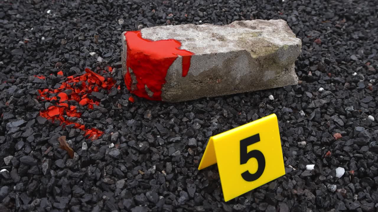 Close up view of a blood covered brick or a blunt force object next to a yellow police crime scene marker on gravel ground where blood spatter and drops are. Shallow depth of field, bokeh background