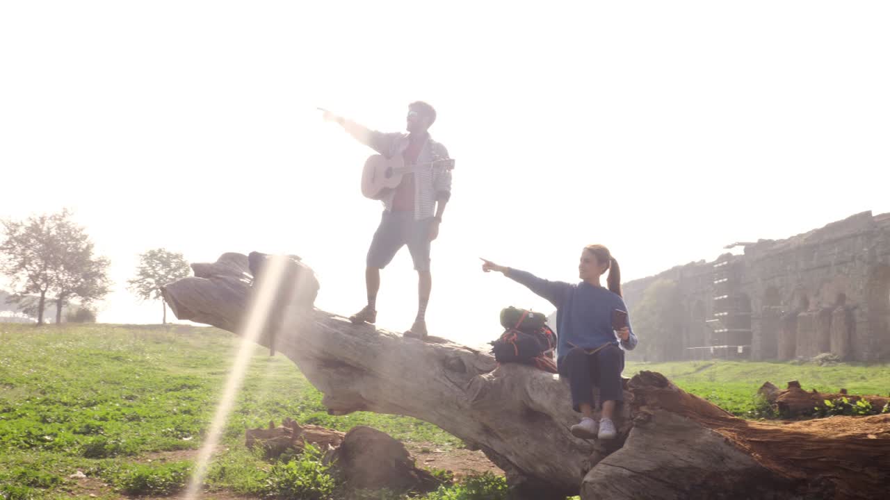 pareja feliz de mochileros turistas en un tronco de tronco señalando dirección con la mano frente a las antiguas ruinas del acueducto romano en el parque degli aquedotti parque en roma al amanecer romántica cámara lenta