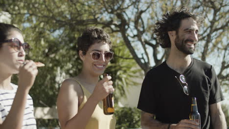 smiling friends talking together while drinking beer and eating pizza on rooftop party