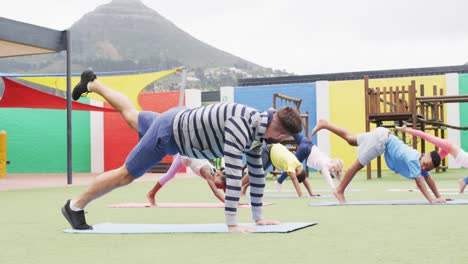 diverse male teacher and happy schoolchildren exercising on mats at school playground