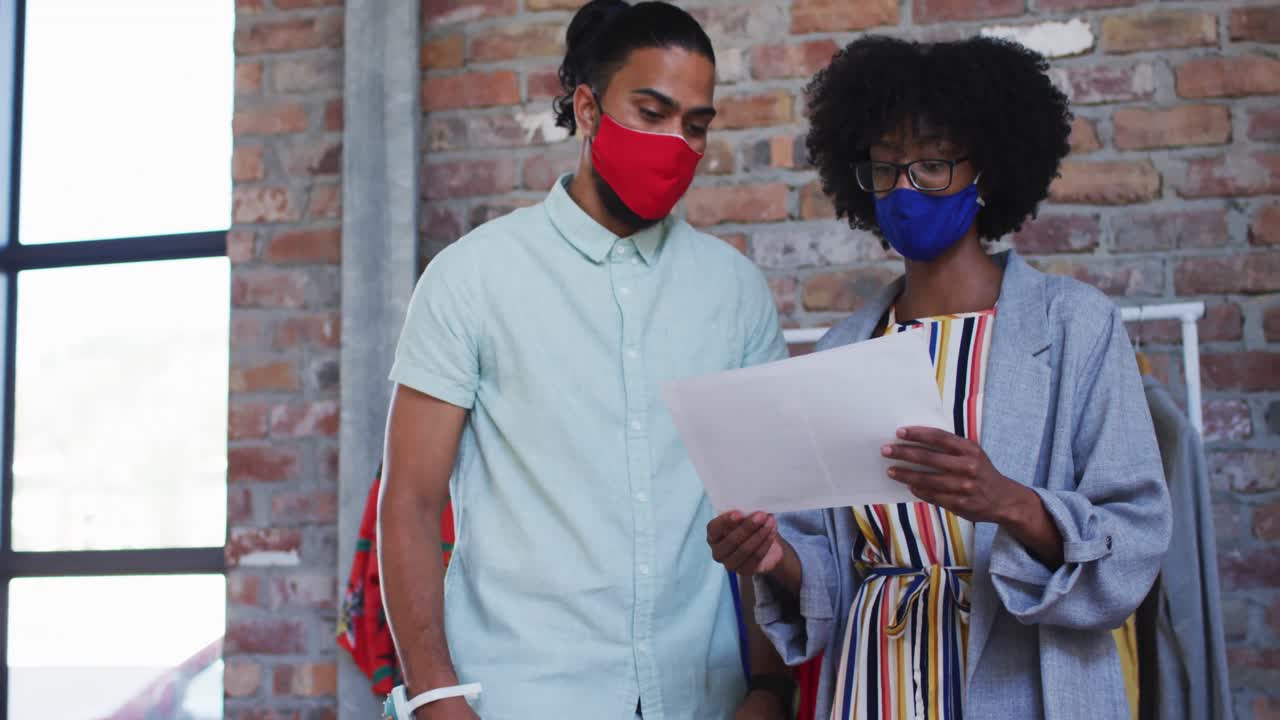 Diverse male and female fashion designers wearing face masks in discussion at work looking at paper
