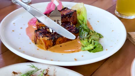 a fork and knife cut into a glazed pork belly steak served with greens and sauce on a white plate, under bright natural lighting