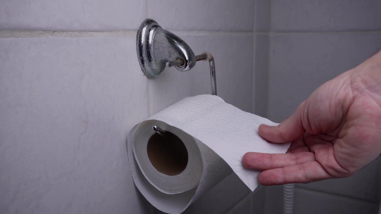 A hand comes into frame and starts touching, rubbing and feeling the fabric of the tissue of a toilet paper roll on a metal holder on the bathroom wall.