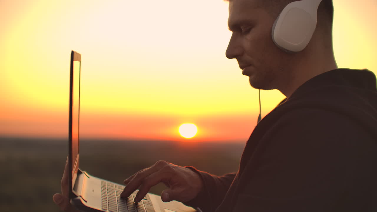 A man in large white headphones types with his fingers on the keyboard of a laptop standing on the roof of a building at sunset against the background of the city.