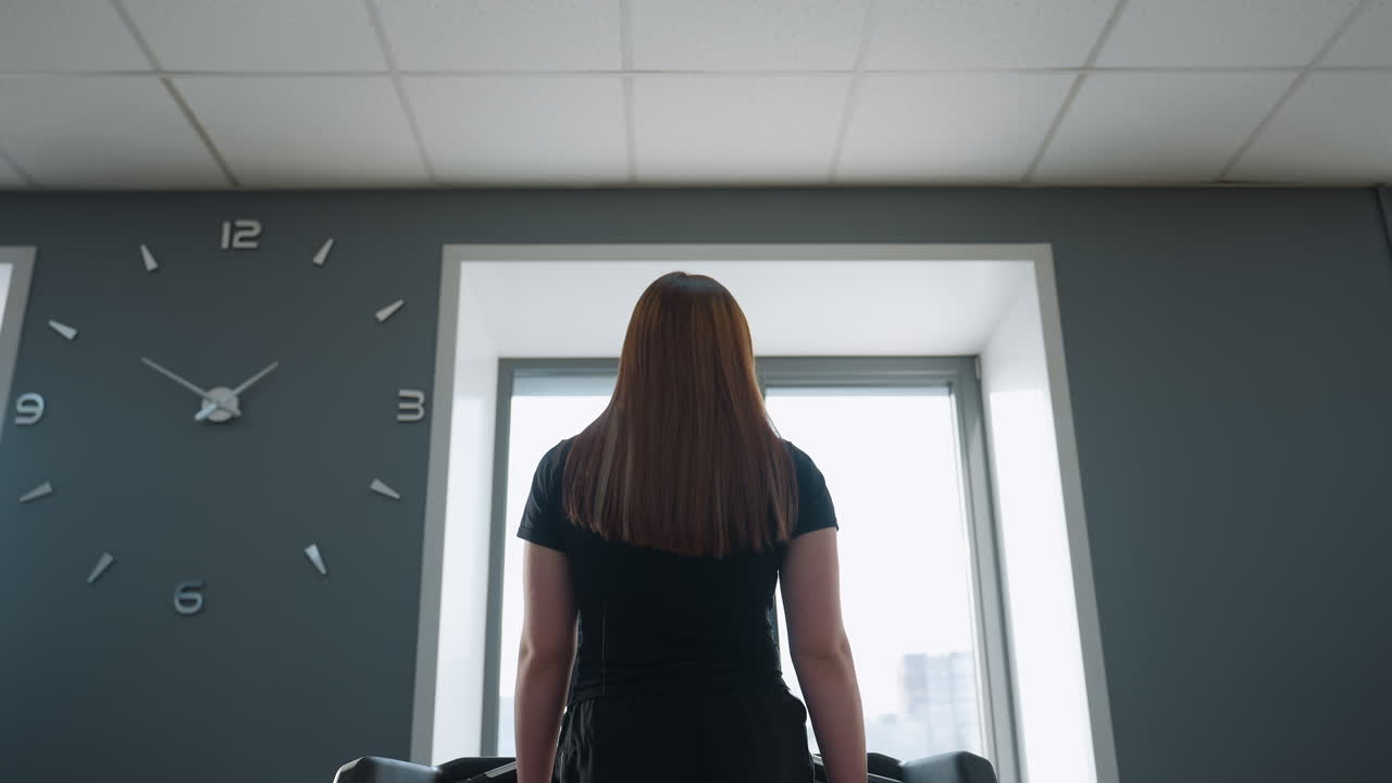 athlete with long brown hair facing large window with bright natural light streaming through, wall clock mounted on gray wall in modern gym interior, standing calmly in workout attire