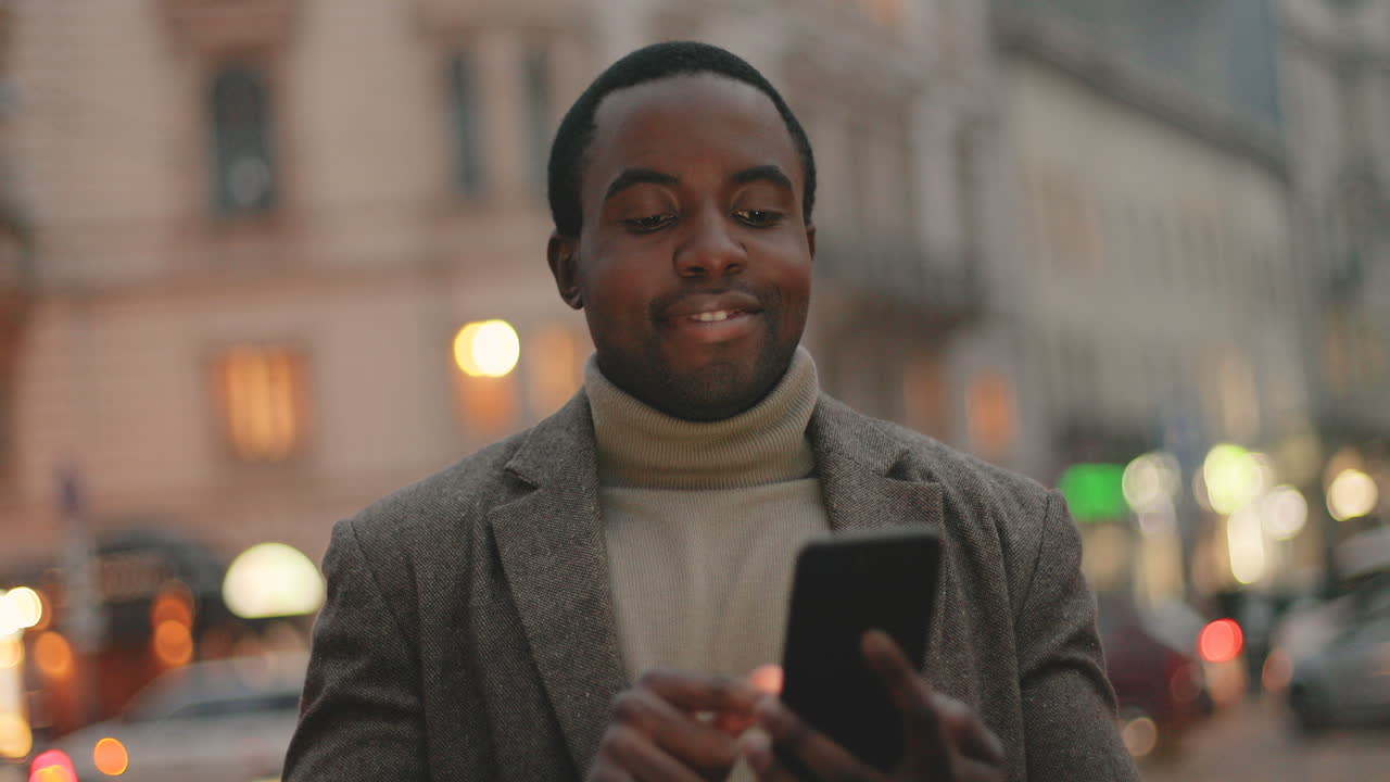 African American businessman wearing coat and texting on his smartphone in the street, then smiles to the camera