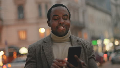 African-American-businessman-wearing-coat-and-texting-on-his-smartphone-in-the-street,-then-smiles-to-the-camera