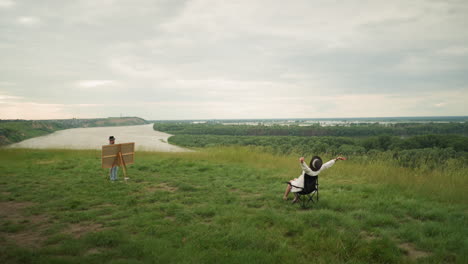 a serene scene capturing a woman in a black hat, sitting comfortably on a chair with her hands touching her ears while an artist paints her in a lush grassy field by a tranquil lake