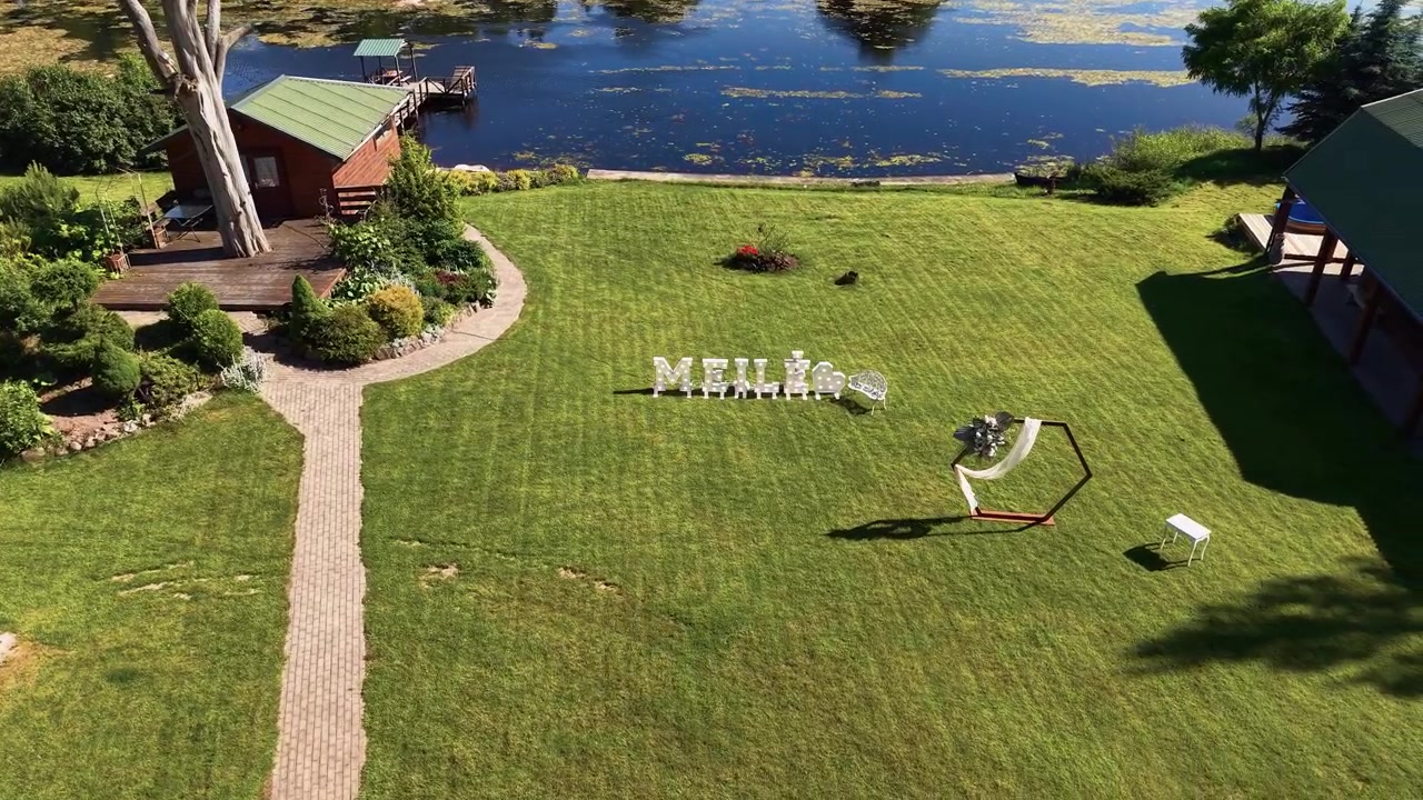 A stunning aerial drone video captures a beautifully landscaped lakeside wedding venue with rustic cabins, lush green grass, decorative arch, and elegant decor under bright daylight.