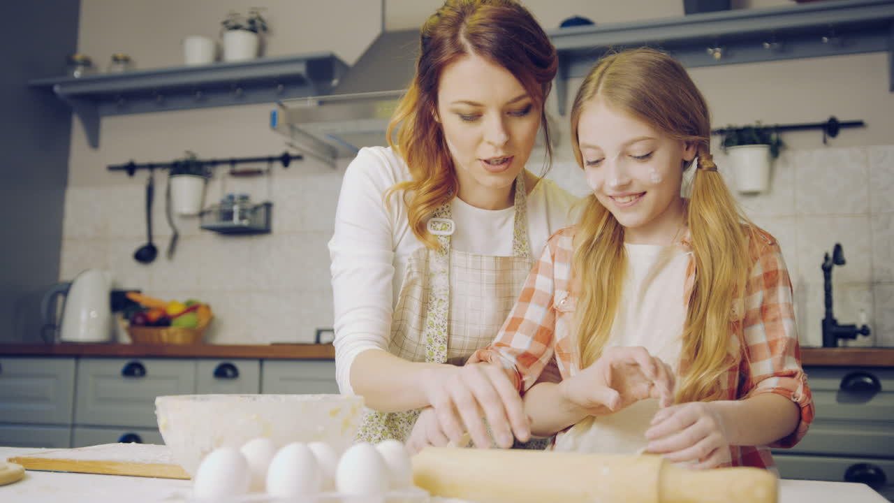 Pretty mother teaching and showing to her young blonde daughter to make a daugh for cookies on the kitchen table in weekend. Indoor