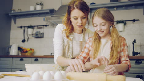 Pretty-mother-teaching-and-showing-to-her-young-blonde-daughter-to-make-a-daugh-for-cookies-on-the-kitchen-table-in-weekend.-Indoor