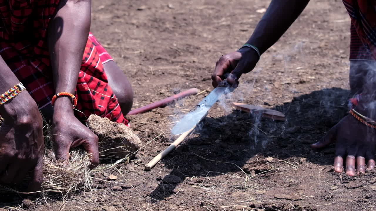 hacer fuego: primer plano de las manos de los maasai creando fuego con un palo y un cuchillo en tierra seca