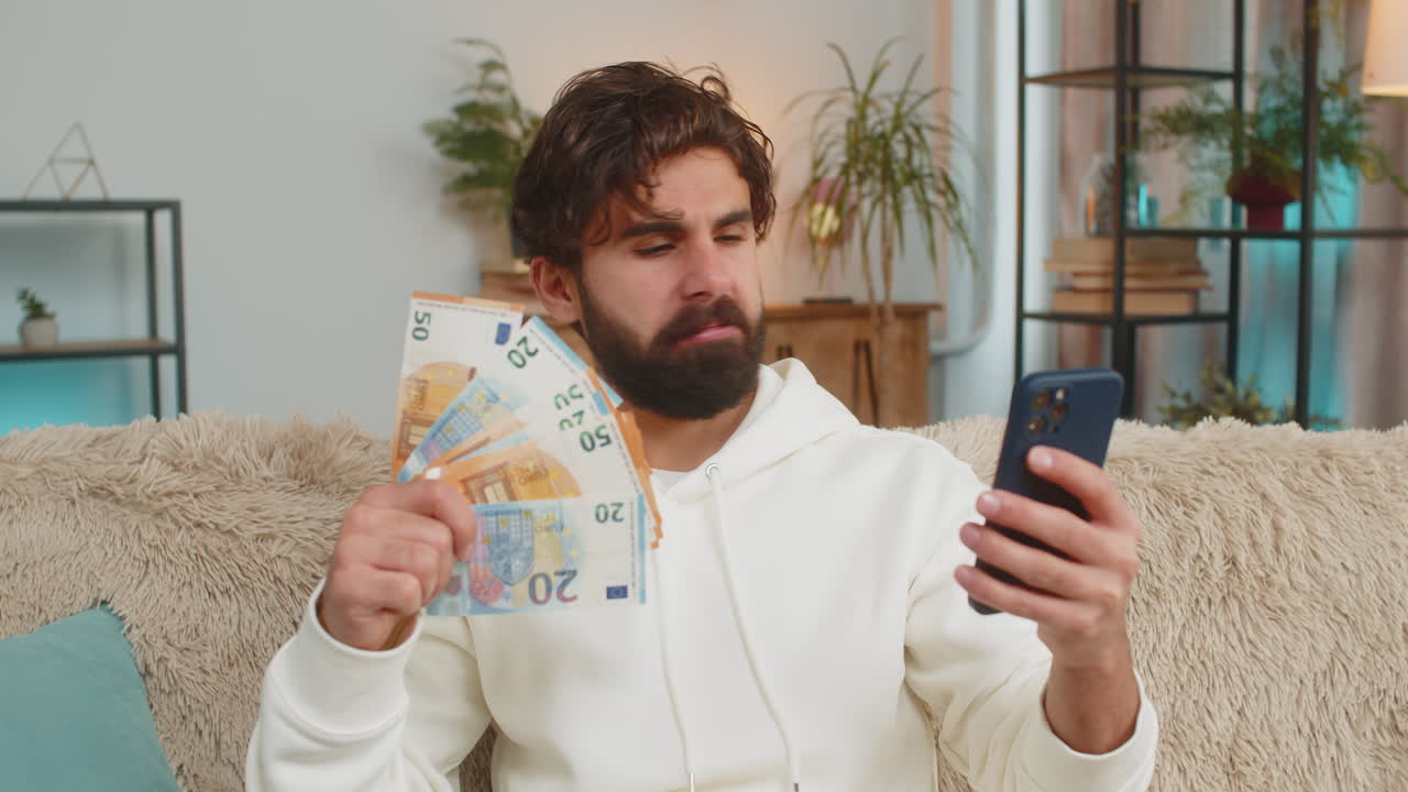 Giovane uomo indiano ricco e di successo che tiene in mano una banconota da un euro e uno smartphone, seduto sul divano di casa.