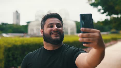 Junger-Bärtiger-Mann-Lächelt-Und-Macht-Ein-Selfie-In-Einem-Botanischen-Garten-Mit-Einem-Großen-Gewächshaus-Im-Hintergrund-In-Curitiba,-Brasilien