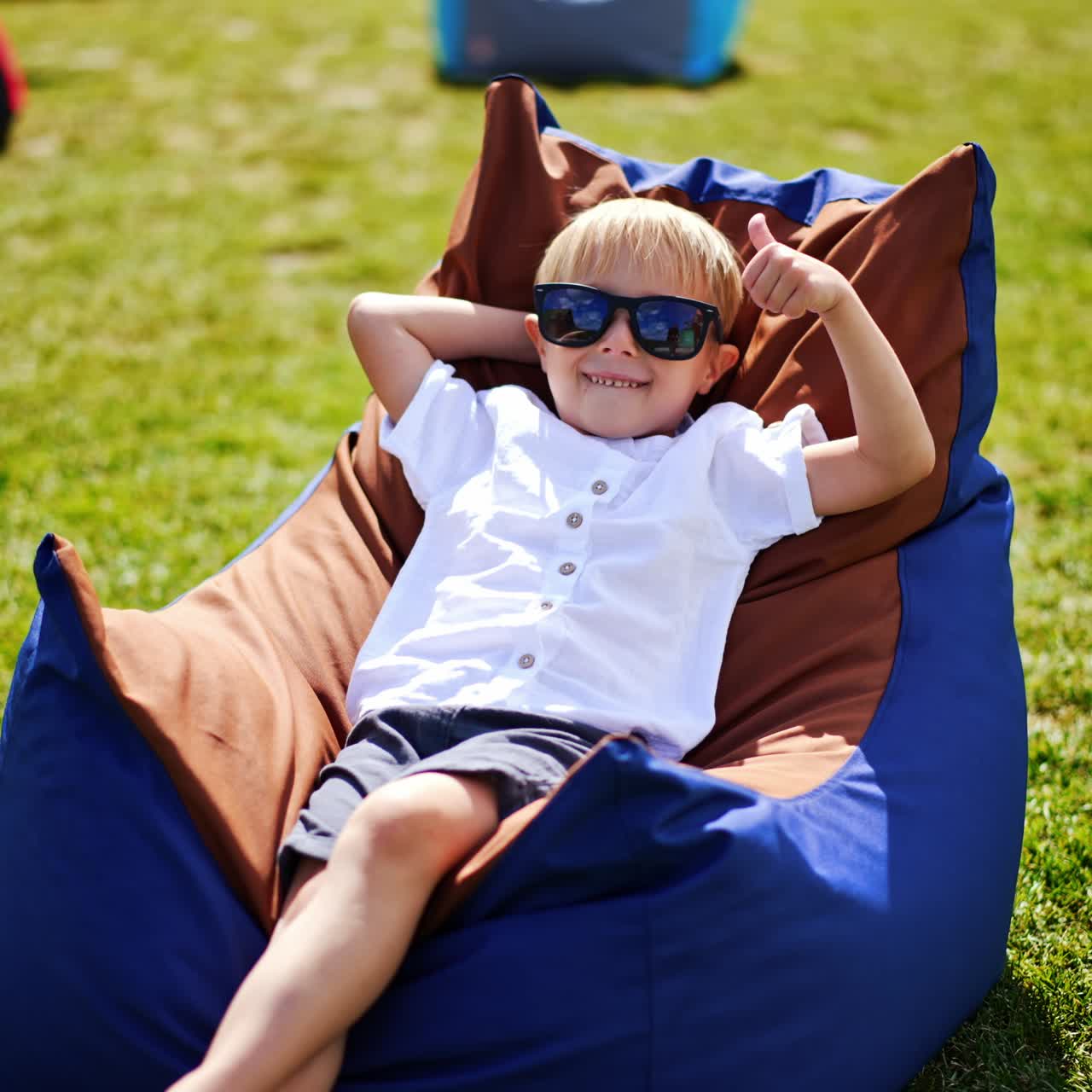 Smiling blond little boy enjoying his time in bean bag chair outdoors. Child in summer clothes and sunglasses relaxing and sunbathing