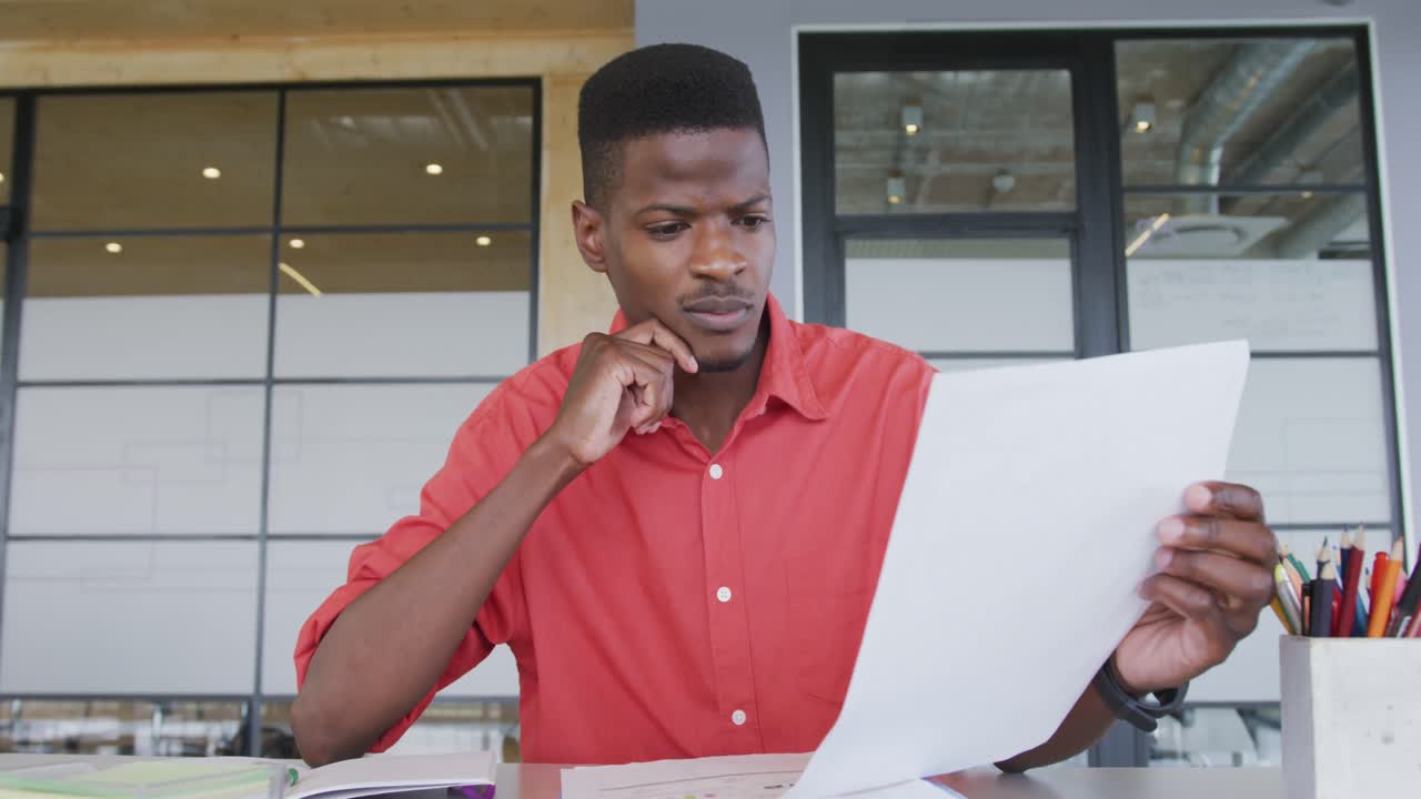 African american creative businessman at desk, reading document and talking during video call