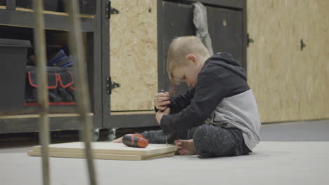 portrait of the blonde boy sitting in the workshop using a screwdriver on wood plank