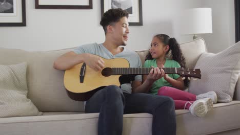 happy biracial father and daughter sitting on sofa playing guitar