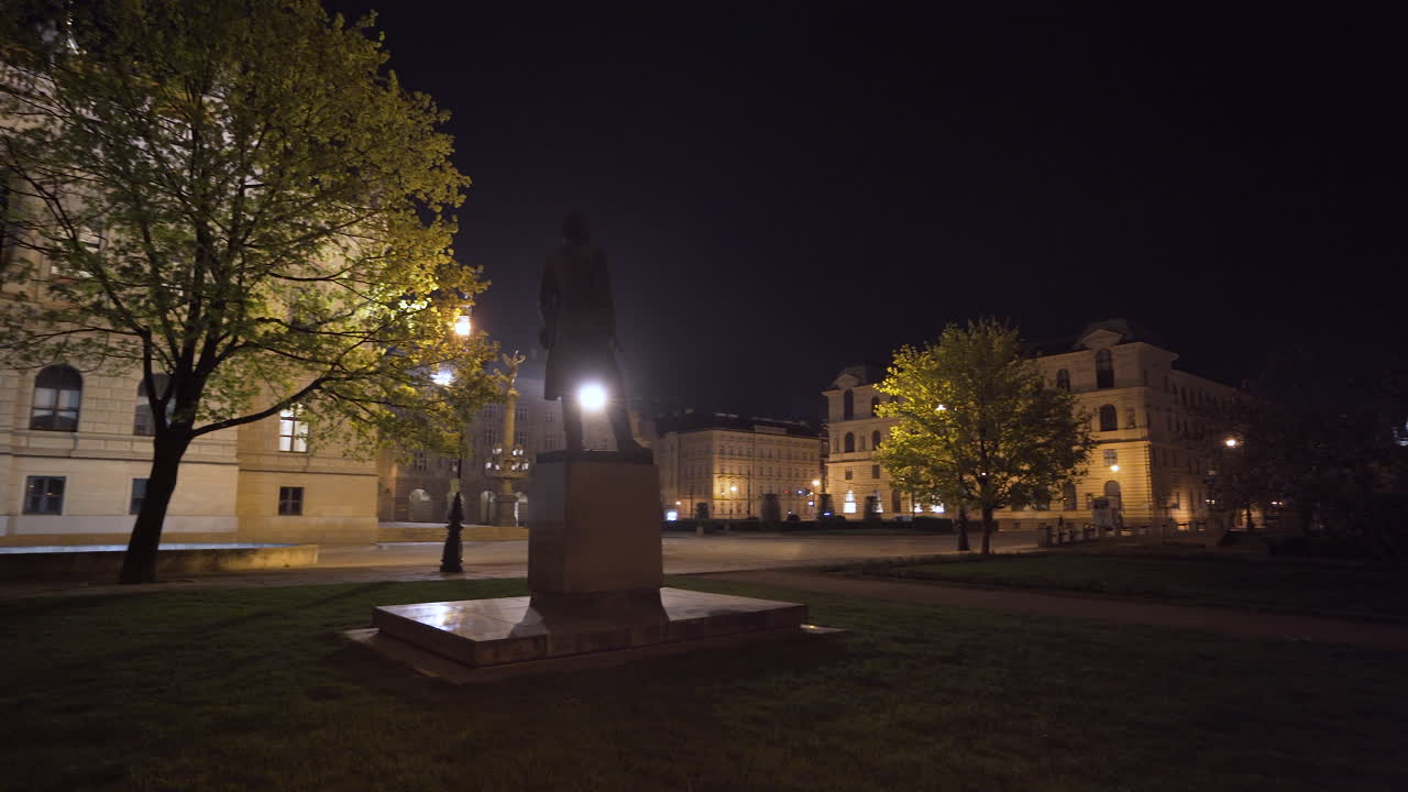 una estatua de una persona en una plaza vacía en praga, chequia, por la noche, durante un cierre covid-19, árboles y la magnífica sala de conciertos de rudolfinum en el fondo, niebla ligera sobre la plaza, pan