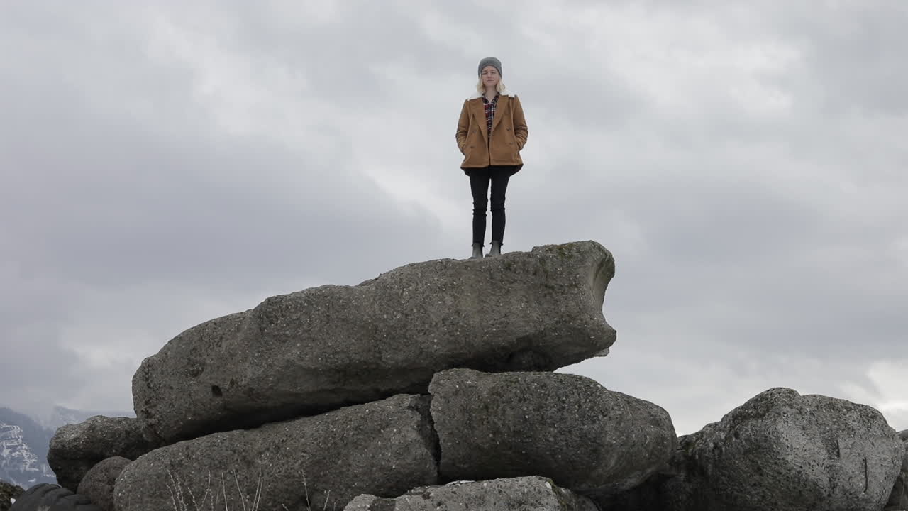 A girl stands on boulders with snowy mountains around her.