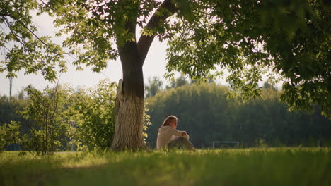 woman sits under large tree with sunlight gently illuminating her, surrounded by lush greenery, background shows distant trees, a football goalpost, and fluttering leaves