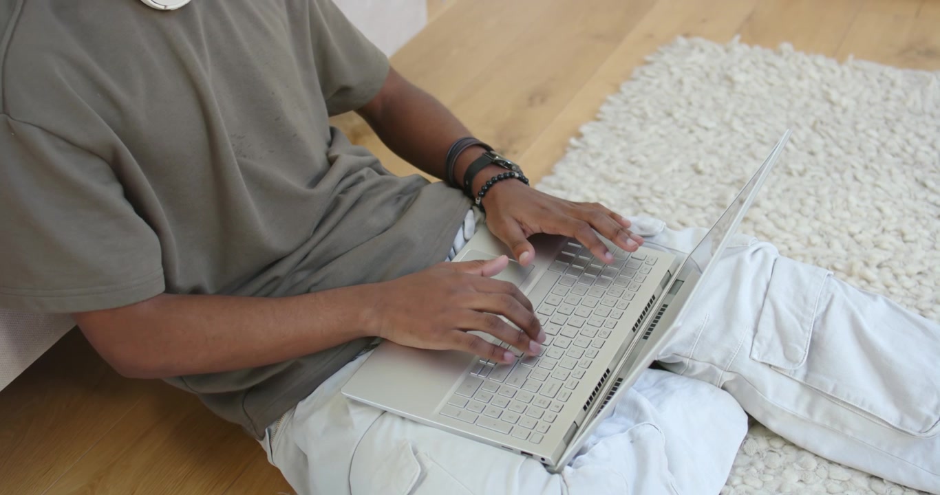 Male sitting on hardwood floor beside white shag rug, positioning hands on laptop, typing for work