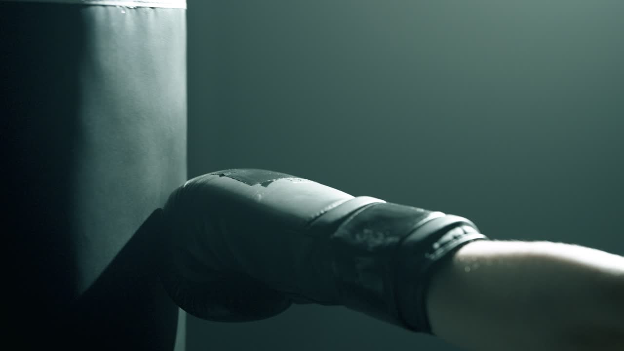 Close-up of blonde woman’s hands in black boxing gloves punching a heavy bag from side profile. Dramatic cinematic light highlights impact and glove texture in a dark, smoky training studio