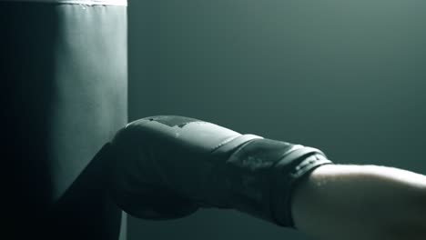 close-up of blonde woman’s hands in black boxing gloves punching a heavy bag from side profile. dramatic cinematic light highlights impact and glove texture in a dark, smoky training studio