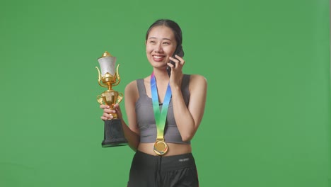 asian woman with a gold medal holding a gold trophy and talking on smartphone on green screen background in the studio