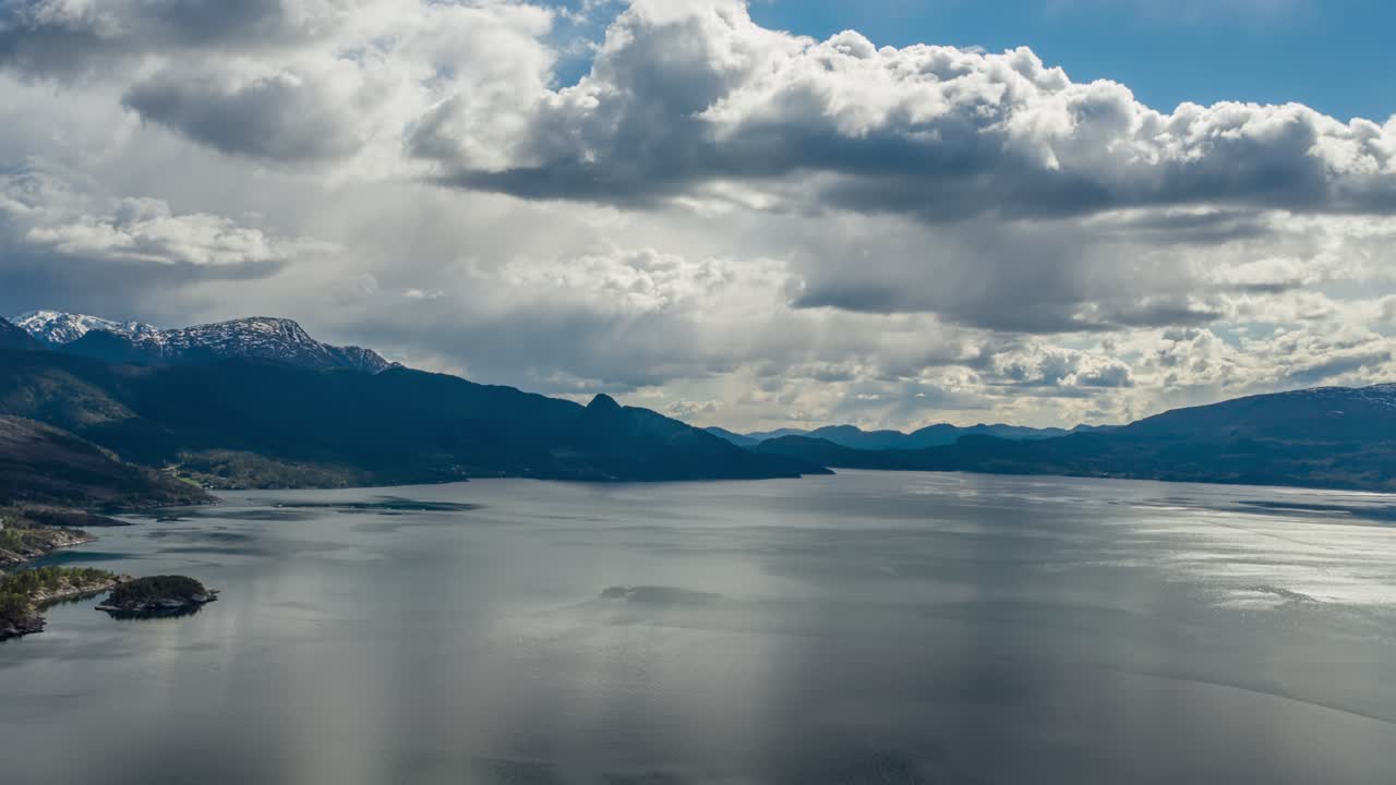 Thick, gray, stormy clouds whirling above the dark blue waters of the Hardanger fjord, Norway. Rays of sun are piercing through the clouds, lighting dark waters. Mountains tower on the horizon