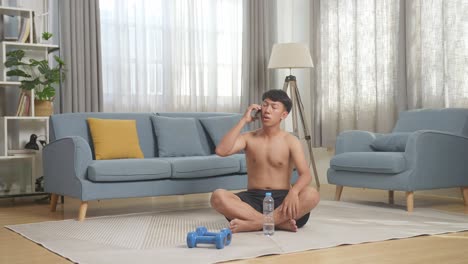 full body of asian teenager boy training with dumbbells on mat and talking on smartphone while doing shirtless workout at home