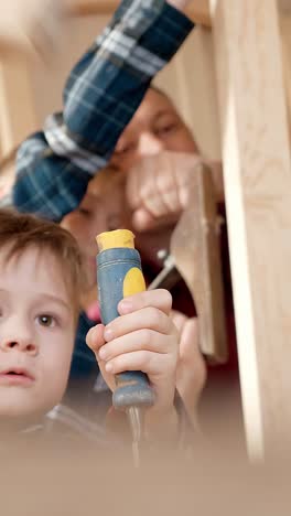 young child is engaged in woodworking activity, holding a tool while an adult demonstrates hammering technique, showcasing hands-on learning and creativity
