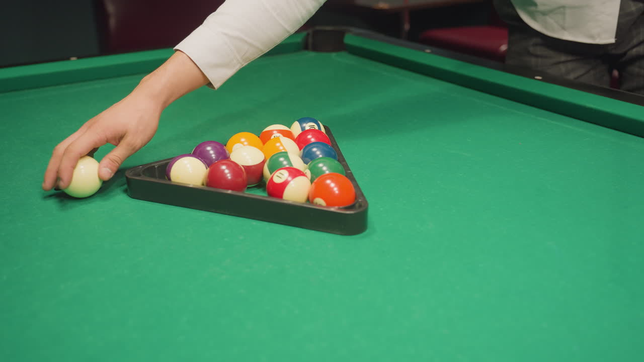 Player hand carefully arranges colorful billiard balls inside triangle rack on green pool table, preparing for break shot in pool hall. Close-up shot captures precise setup before game begins