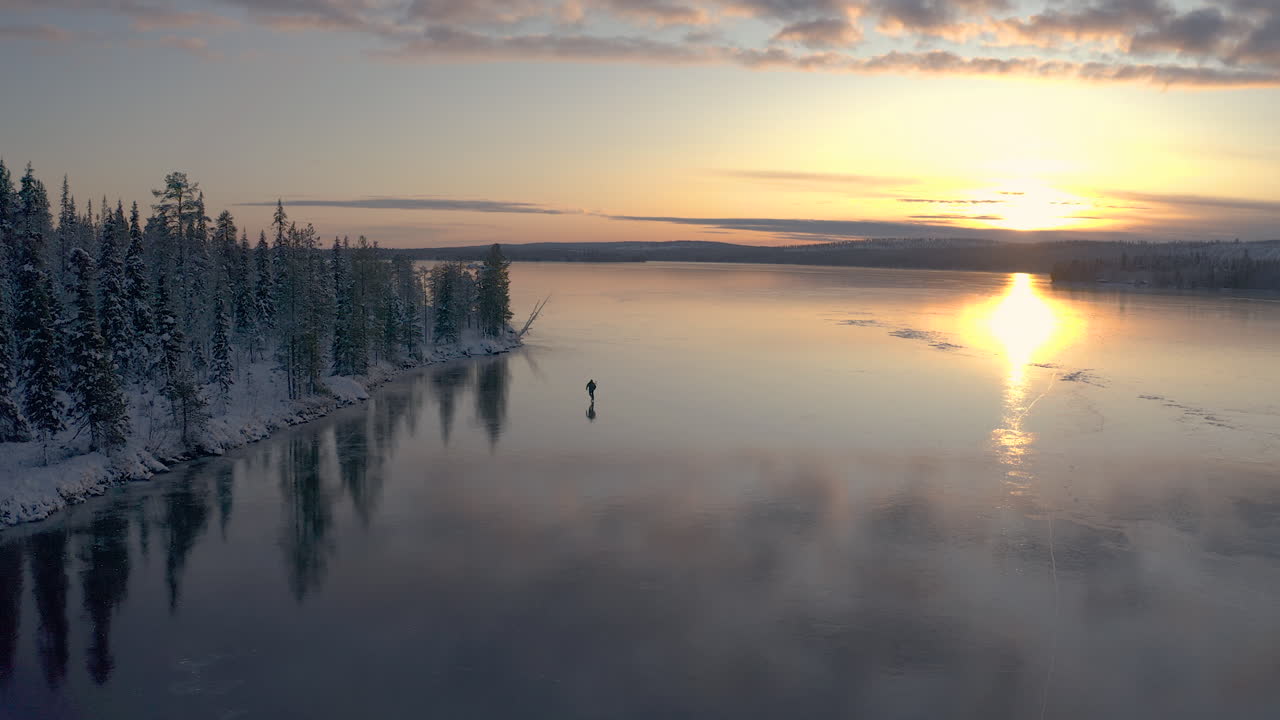siguiendo a un patinador de hielo nórdico en un lago congelado desde una vista aérea de ensueño mientras el patinador se desliza a través del hielo limpio junto a un bosque invernal nevado y la puesta de sol tiñe el fondo de naranja