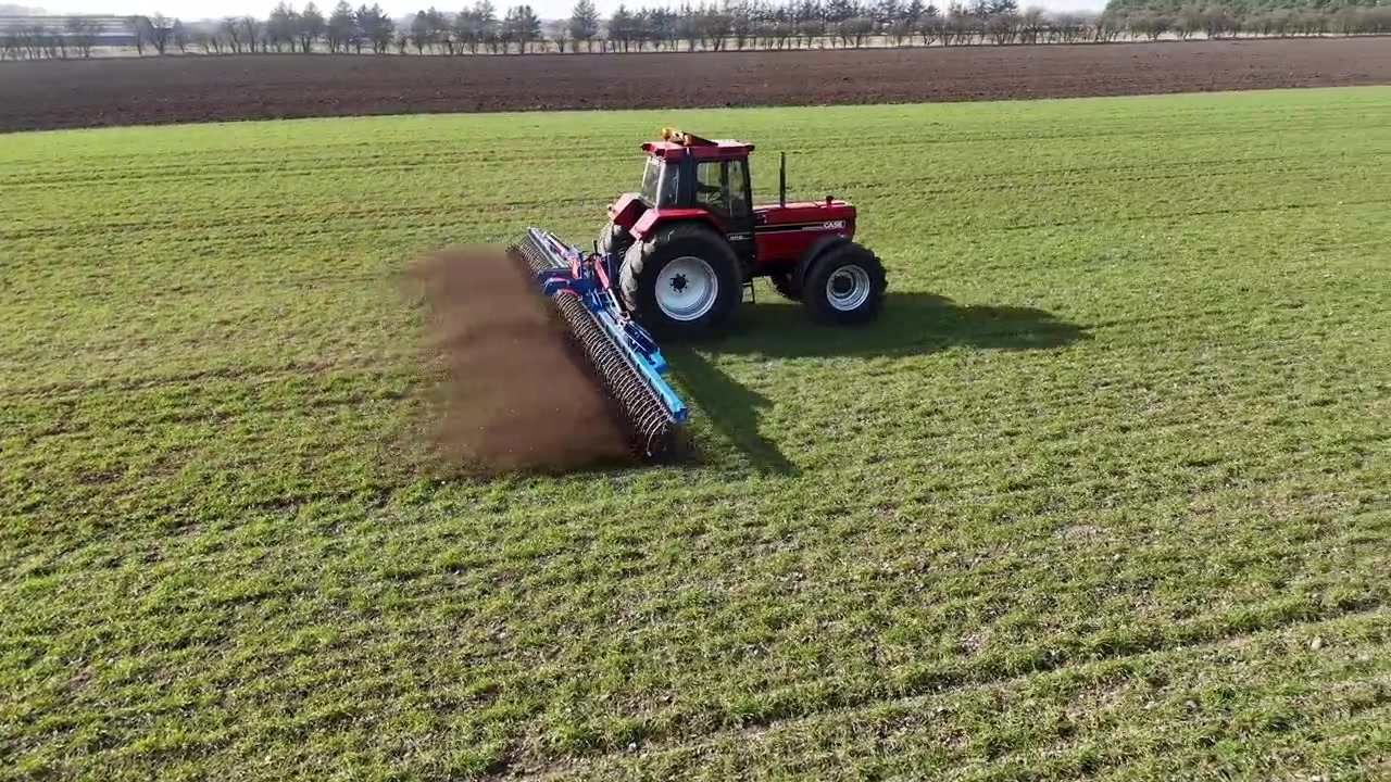 Un'inquadratura cinematografica con drone segue un trattore rosso che ara un campo agricolo verde e rivela l'aratro in funzione dietro di esso
