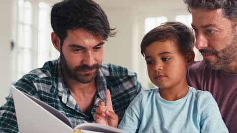 close up of same sex family with two dads and son reading book in kitchen at home together