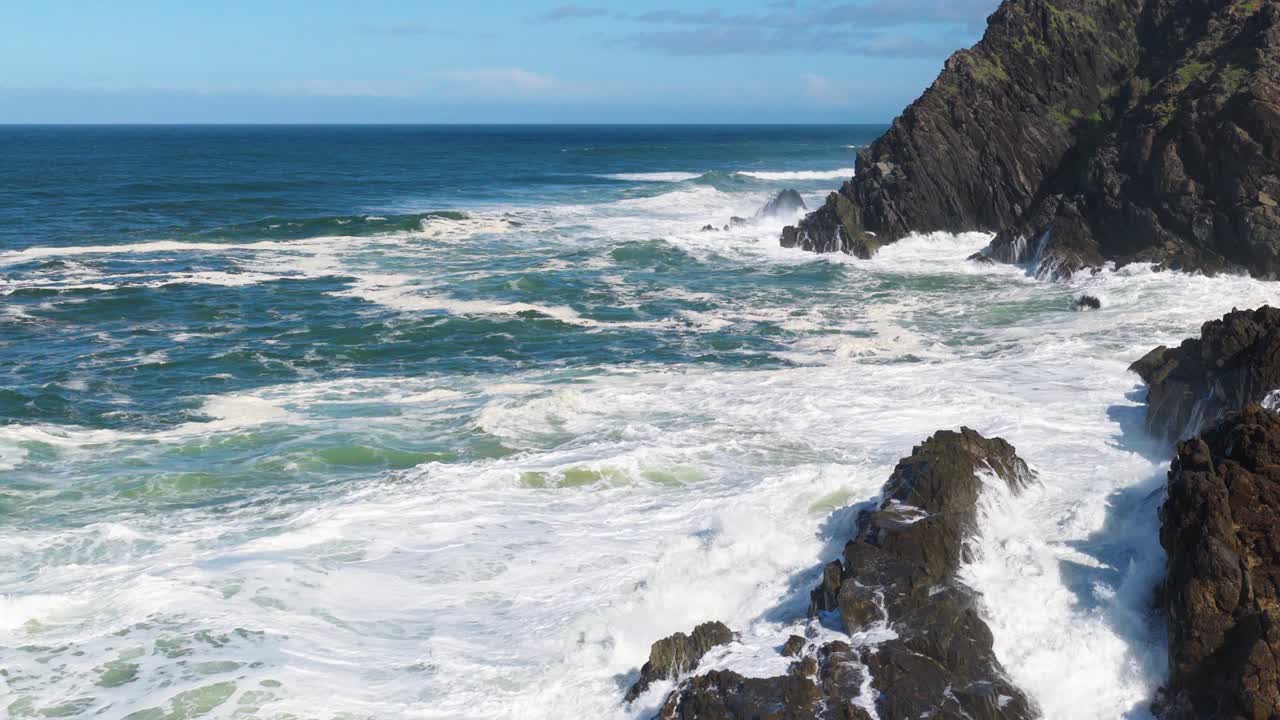 Dynamic ocean waves crash against rocky cliffs at Byron Bay, under clear skies and bright sunlight, creating a dramatic seascape