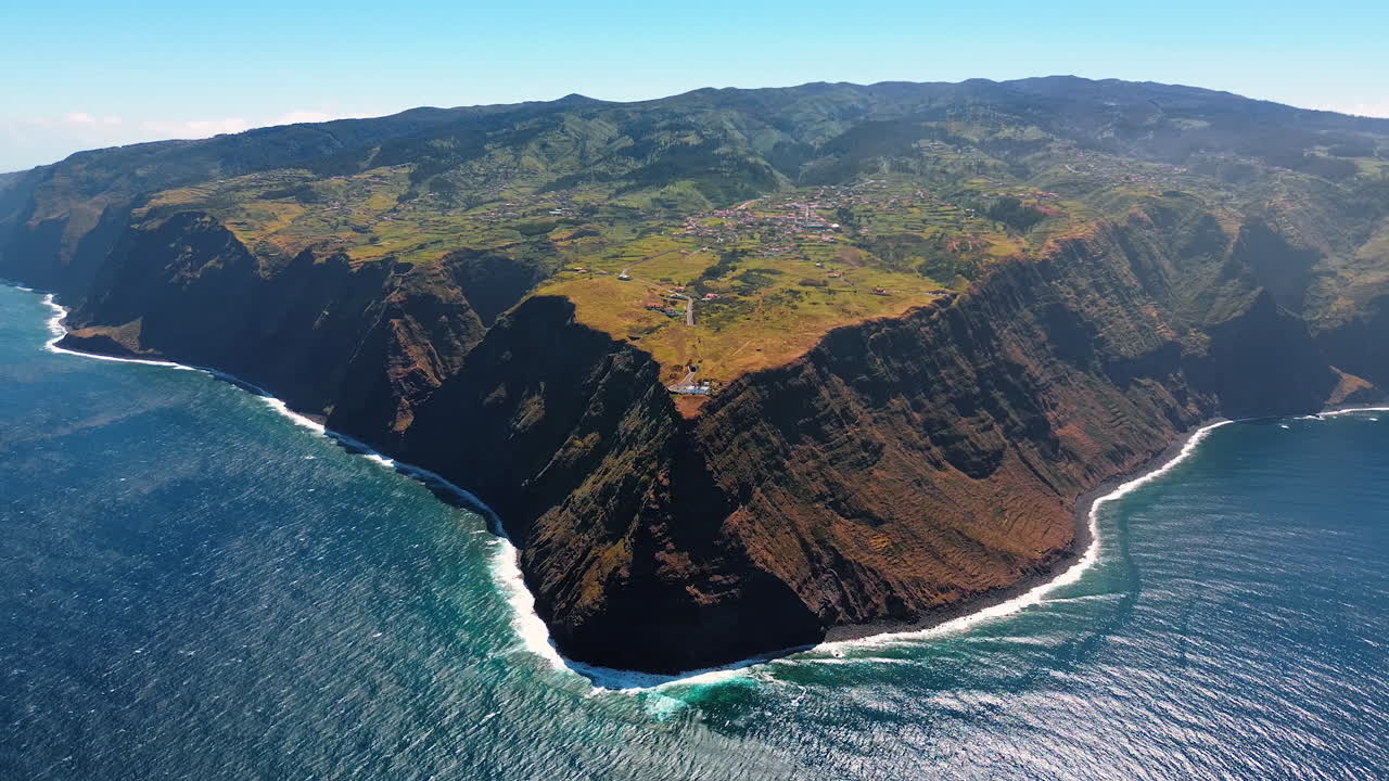 Triangular high rock deeps into the blue waterscape of the Atlantic Ocean. A town is located on the top of the mountain. Top view on the Madeira Islands, Portugal.