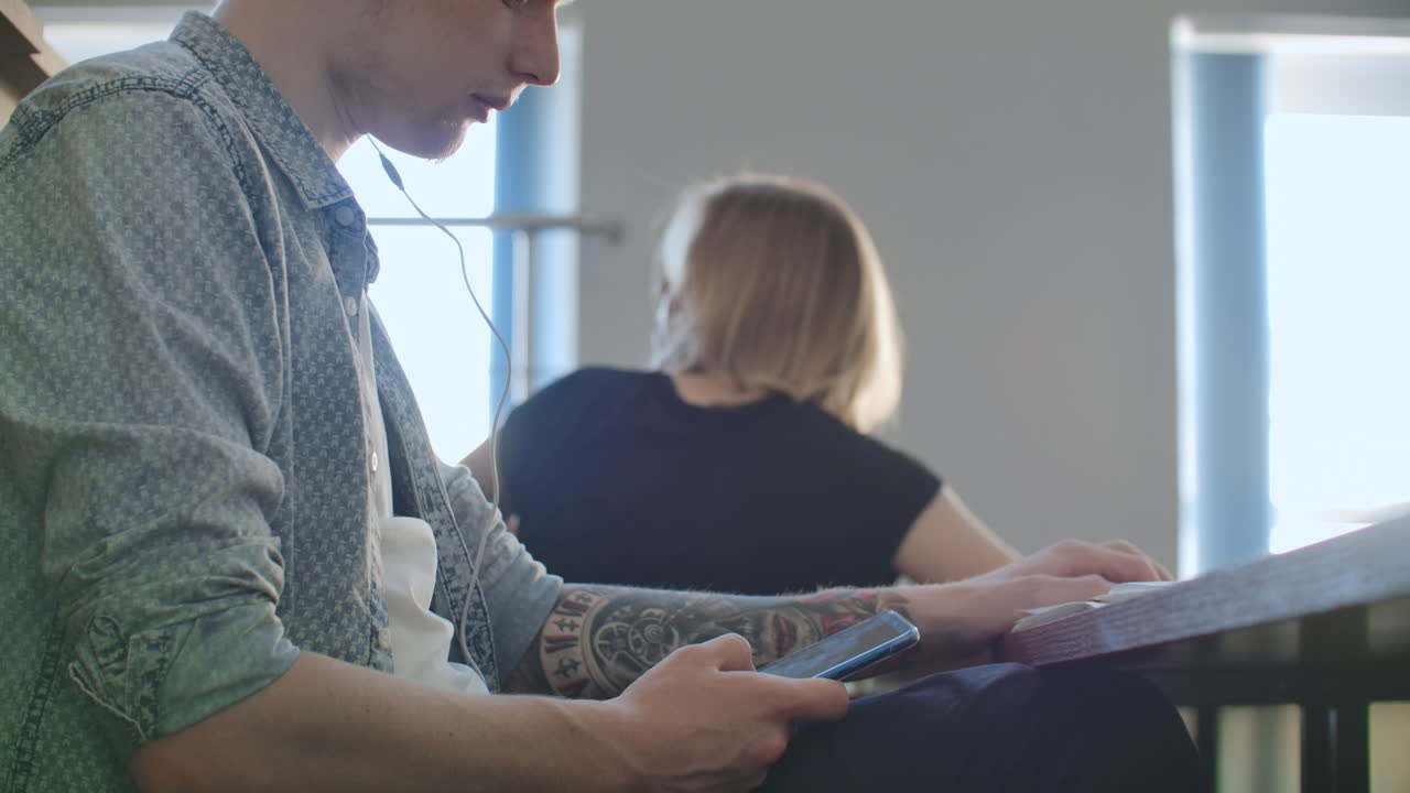 un estudiante masculino en un auditorio de la universidad escucha música con auriculares blancos durante un descanso sin prestar atención a los demás. mucha gente en la audiencia está hablando.