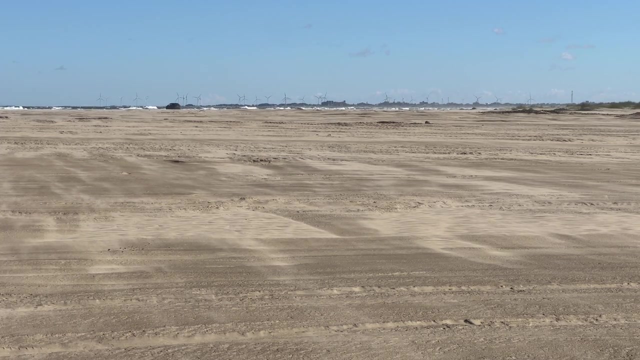 Statisk udsigt til vind, der skaber flydende sandmønstre på en flad strand nær en fjern vindmøllepark.