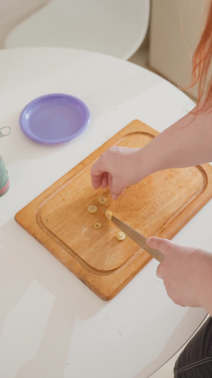 Una vista ravvicinata dall'alto mostra mani che affettano un piccolo ingrediente in pezzi sottili su un tagliere di legno caldo accanto a un tavolo rotondo bianco e un piatto color lavanda, con un attento movimento del coltello verso il basso in tranquillità.