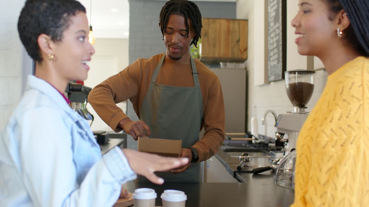 Female friends gesturing confirming order, barista packing coffee cups, pastry box at cafe counter