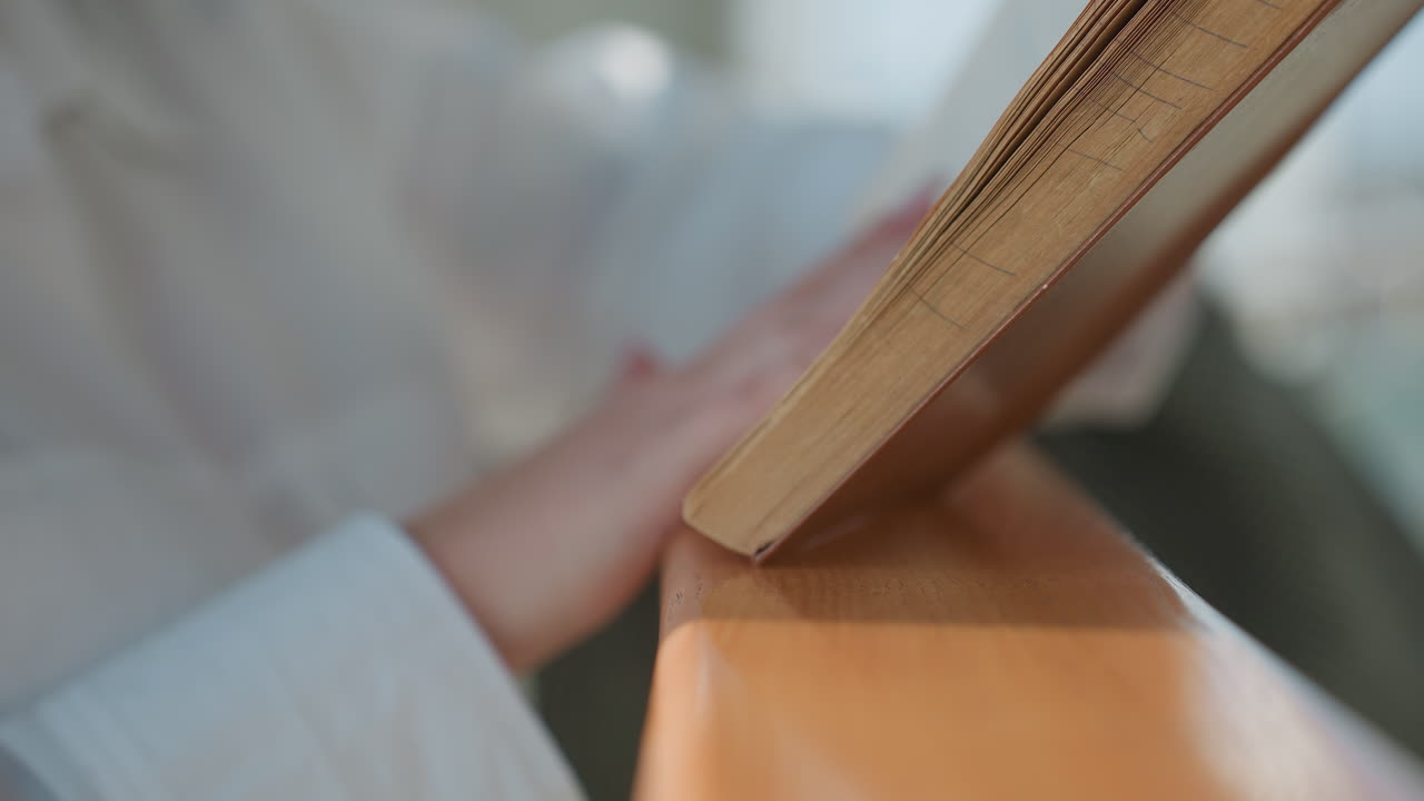 Young woman with manicured nails flips pages of paperback novel while resting book on wooden railing in sunlit modern interior setting, showing delicate interaction between hand