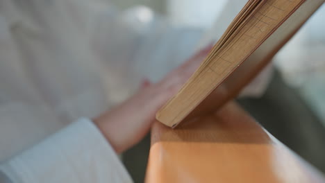 young woman with manicured nails flips pages of paperback novel while resting book on wooden railing in sunlit modern interior setting, showing delicate interaction between hand